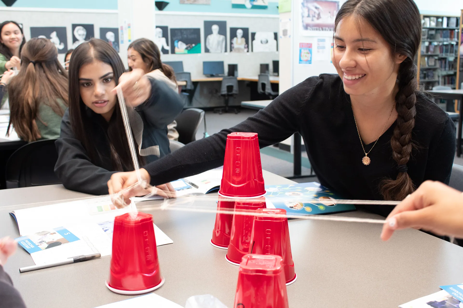Young women doing an activity with plastic cups and rubber bands on table